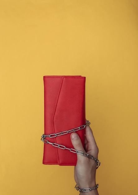 Hand wrapped in a steel chain holds a purse on yellow background.