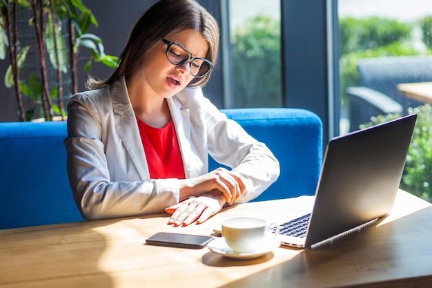 Hand wrist pain. Portrait of sad stylish brunette young woman in glasses sitting holding her painful hand after long time working and typing on laptop. indoor studio shot, cafe, office background.