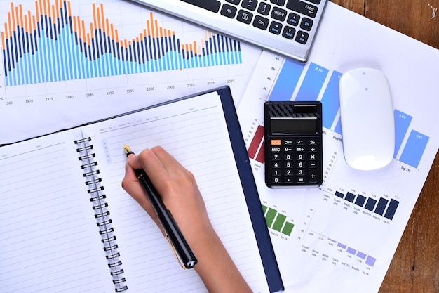 Hand writing on blank notebook with graph, chart, keyboard, mouse and calculator on wooden table