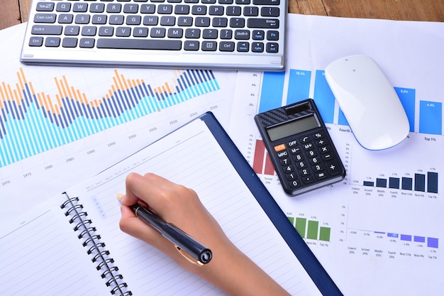 Hand writing on blank notebook with graph, chart, keyboard, mouse and calculator on wooden table