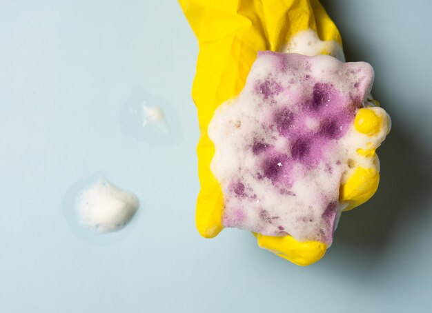 A hand in a yellow rubber glove holds a cleaning sponge in\
soapy foam on a blue background. house cleaning concept, cleaning\
service