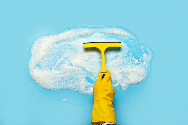 Hand in a yellow rubber glove holds a scraper for cleaning and wipes soap suds on a blue surface. Cleaning concept, cleaning service. . Flat lay, top view