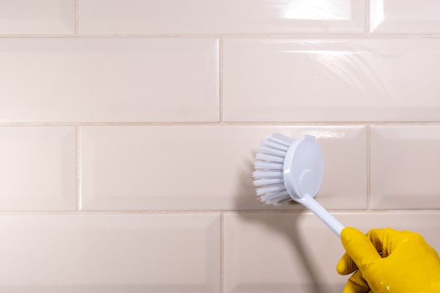 Hand in a yellow rubber glove wiping the ceramic tile surface with a white brush, close-up, copy space. Removal of stains, disinfection of the kitchen, bathroom. House cleaning concept