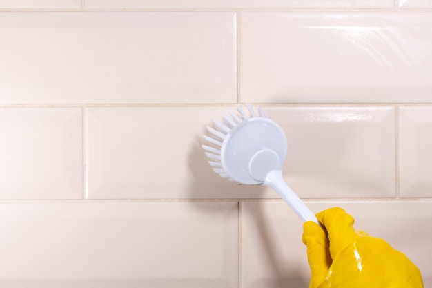 Hand in a yellow rubber glove wiping the ceramic tile surface with a white brush, close-up, copy space. Removal of stains, disinfection of the kitchen, bathroom. House cleaning concept