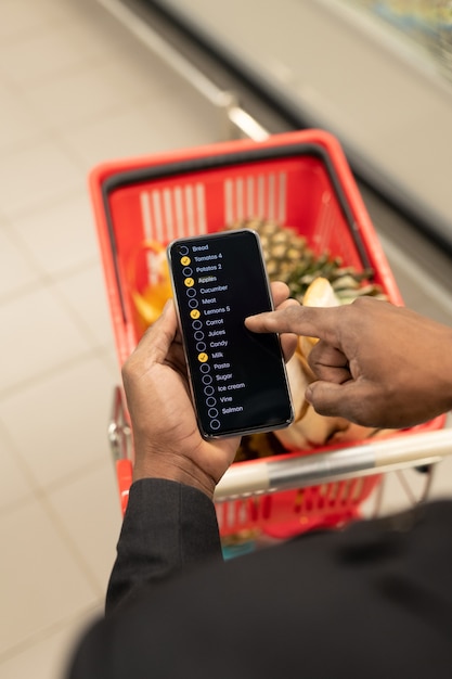 Hand of young African man with smartphone pointing at list of stuff to buy while pushing shopping cart with goods in modern supermarket