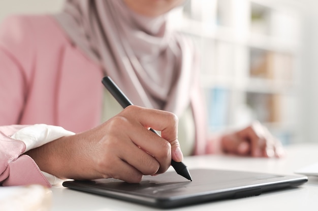 Mano di giovane designer femminile contemporanea in hijab che tiene lo stilo sul tablet mentre ritocca le immagini o disegna uno schizzo dalla scrivania in ufficio