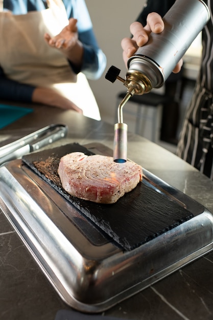 Photo hand of young cooking coach in apron smoking or processing piece of boiled beef on special board by table with one of trainees near by