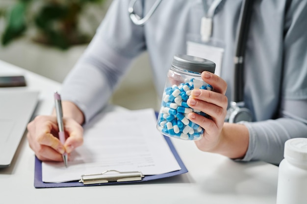 Foto mano di giovane medico femminile che tiene il barattolo con le pillole e che fa la prescrizione