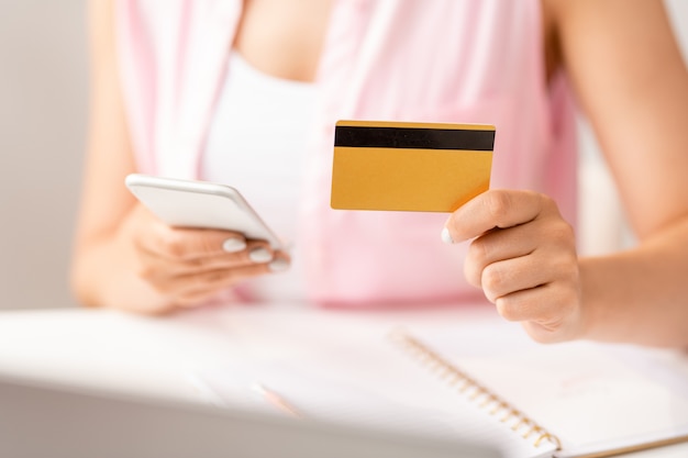 オンライン注文をするプロモーションを探しているスマートフォンを使用してプラスチックカードを持つ若い女性の買い物客の手