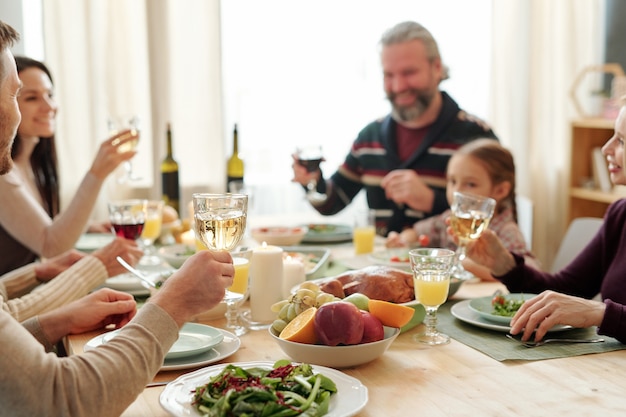 Foto mano del giovane che tiene un bicchiere di vino sul tavolo servito durante il brindisi alla cena di famiglia festosa il giorno del ringraziamento