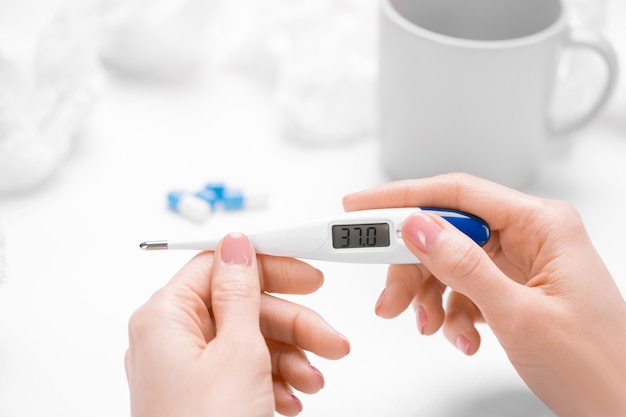 Hand of young sick female holding thermometer showing temperature of 37 degrees over table with mug, pills and crumpled tissues