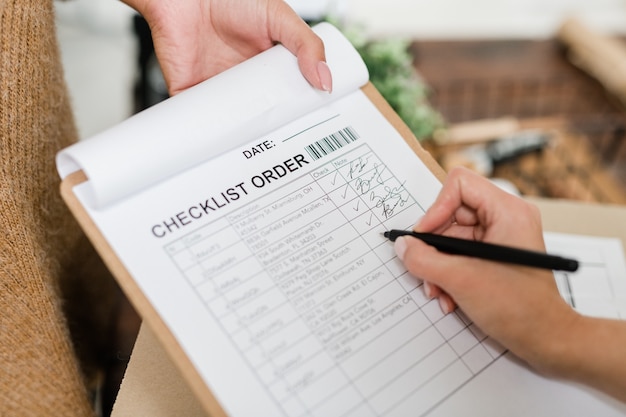 Hand of young woman with pen going to put tick opposite one of positions in checklist after receiving order