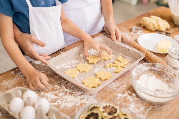 hand of youngster putting raw biscuits on tray while helping mom with dough and cookies by kitchen table
