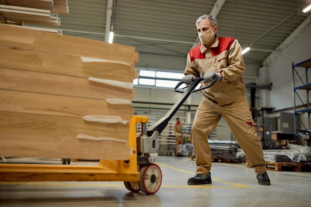 Handarbeider met gezichtsmasker die palletkrik met houten planken duwt bij timmerwerkplaats