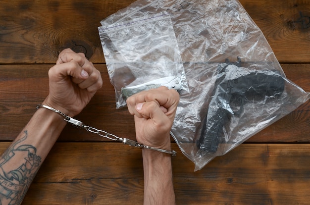 Handcuffed hands of criminal suspect on wooden table and handgun with jackknife in transparent plastic packs