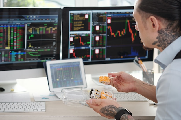 Handelaar die sushi eet voor de lunch en beursgegevens in de gaten houdt op veel computerschermen in zijn kantoor