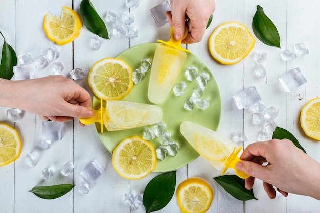 Handen die citroenijslollies oppakken van een bord op een witte houten tafel bedekt met ijsblokjes, schijfjes citroen en natuurbladeren