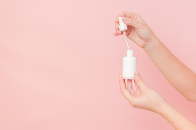 Handen die een glazen fles met een druppeldop vasthouden Witte container met een cosmetisch product serum essentiële olie op roze achtergrond Concept van schoonheid