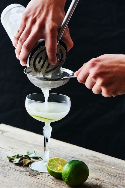 Handen gieten door een zeef in een glazen cocktail Daiquiri op een houten tafel op zwarte achtergrond