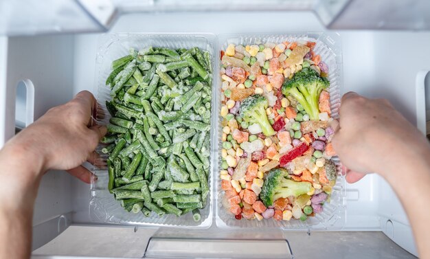 Handen halen een bakje met diepvriesgroenten uit de vriezer van de koelkast