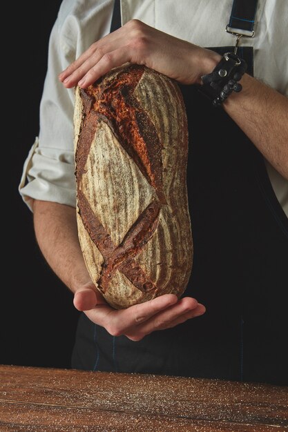 Handen houden het verse brood van een donker schort en een houten tafel op de zwarte achtergrond