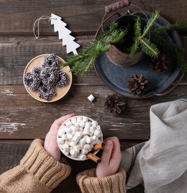 Handen houden kerstmok cacao en marshmallow met kaneel op een oude bruine houten tafel met koekjes, kegels en spar. Nieuwjaar stemming. Bovenaanzicht