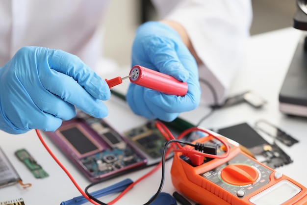 Handen in blauwe handschoenen houden een rode lithium-titaniumbatterij en een voltmeter vast, close-up. Indicator batterijcapaciteit, opladen