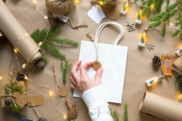 Handen in een witte trui verpakken een cadeau voor Kerstmis en Nieuwjaar in milieuvriendelijke materialen: kraftpapier, levende dennentakken, kegels, touw. Tags met mock up, natuurlijk decor, handgemaakt, DIY. Feestelijke stemming