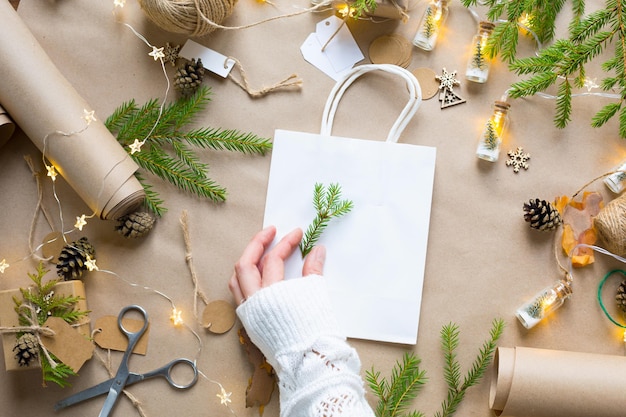 Handen in een witte trui verpakken een cadeau voor Kerstmis en Nieuwjaar in milieuvriendelijke materialen: kraftpapier, levende dennentakken, kegels, touw. Tags met mock up, natuurlijk decor, handgemaakt, DIY. Feestelijke stemming