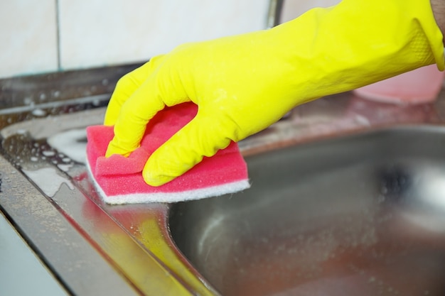 Handen in gele handschoenen schone gootsteen met spons en een speciaal reinigingsmiddel, wasmiddelspray. Hygiëne in de keuken, schoonmaken van de keuken, huishouden.