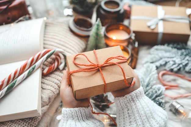 Handen in gezellige trui opening kerstcadeau met rode strik op de achtergrond van de kerstboom met verlichting Stijlvolle vrouwelijke holding aanwezig met rood lint in feestelijke kamer close-up Vrolijk kerstfeest