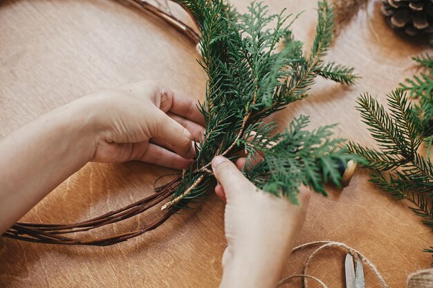 Foto handen met cedertakken en dennenappels draadschaar op houten tafel details voor workshop kerstkrans maken rustieke kerstkrans maken