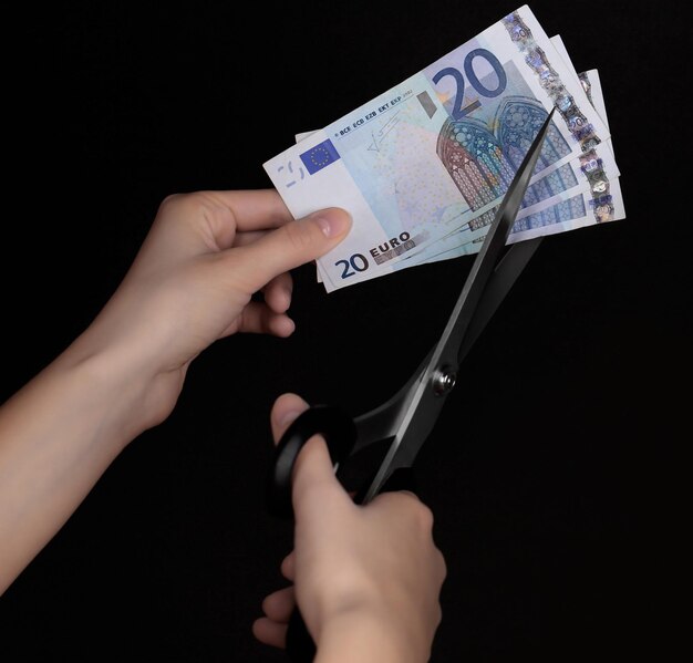 Handen met een schaar die eurobankbiljetten op zwarte achtergrond knippen