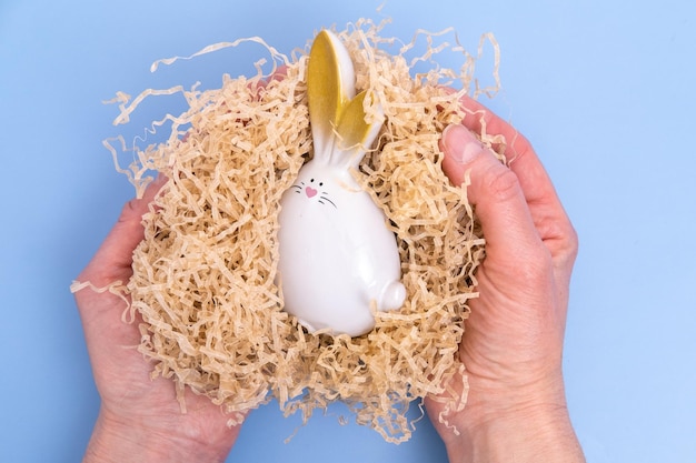 Handen met een witte paashaas zittend in een zelfgemaakte nest op een blauwe achtergrond Pasen minimale creatieve samenstelling