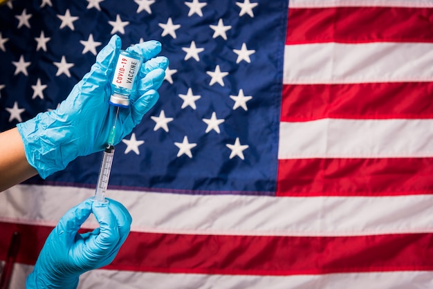 Handen van arts dragen handschoenen met coronavirus COVID-injectieflaconvaccin en spuit op vlag Verenigde Staten van Amerika