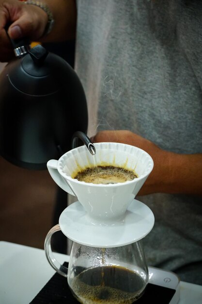 Handen van barista die een infuus hete espresso zet door heet water in een witte kop te gieten met filtreerpapier druppel koffie uit zwarte ketel op witte tafel close-up
