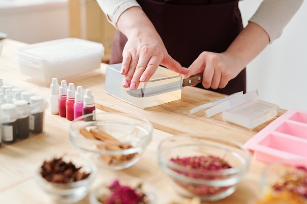 Handen van craftswoman met mes snijden stuk van grote stuk harde zeep massa op houten bord tijdens het werken in de studio