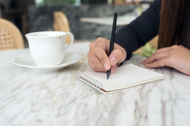 handen van de vrouw te neerschrijven op een witte lege notitieblok met koffiekopje en dessert op tafel