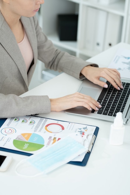Handen van een jonge hedendaagse zakenvrouw of secretaresse in een elegant grijs pak die op de toetsen van het toetsenbord van de laptop drukt tijdens het netwerken op kantoor