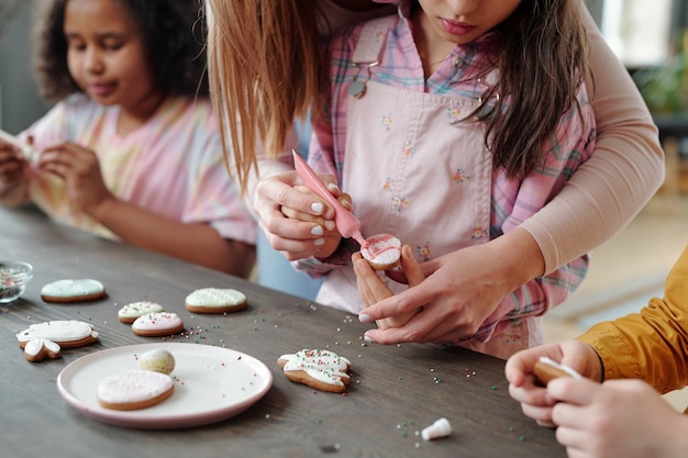 Handen van een jonge vrouw die haar dochtertje helpt met het glazuren van verse zelfgemaakte koekjes of peperkoek terwijl ze allebei aan tafel staan