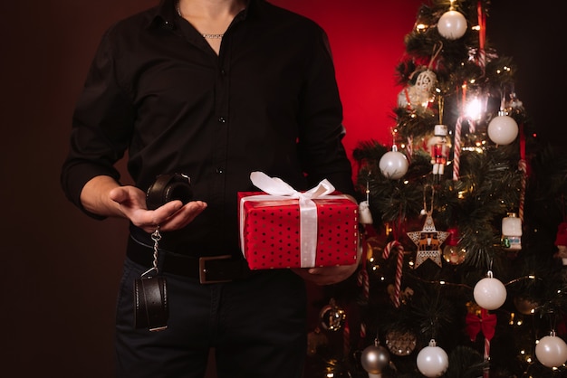 Handen van een man met een geschenkdoos en BDSM handboeien bij de kerstboom. Erotisch cadeau voor het nieuwe jaar voor seksuele spelletjes