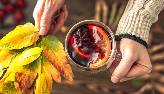 Handen van een vrouw in een trui houden een glazen beker met glühwein en een boeket gele herfstbladeren Concept van een traditionele verwarmende drank