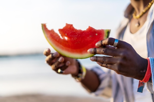 Handen van een zwarte etnische man genieten van de zomervakantie op het strand en eten een watermeloen