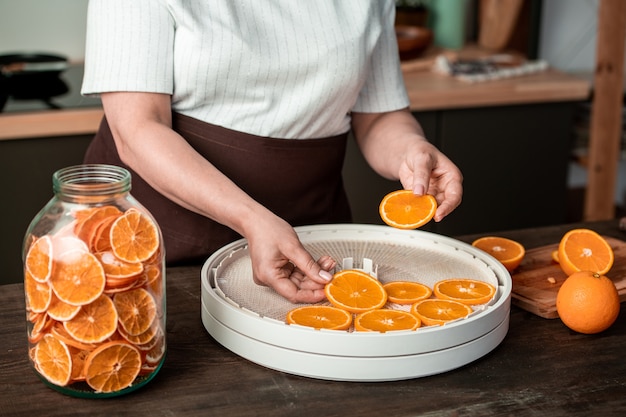 Handen van huisvrouw stukjes sinaasappel op plastic bakjes van voedseldroger zetten tijdens het maken van gekonfijte citrusvruchten voor de winter in de keuken