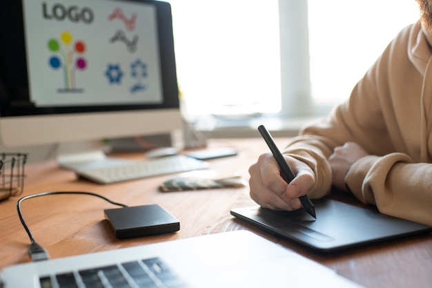 Handen van jonge creatieve mannelijke freelance ontwerper die touchpad en stylus gebruikt tijdens het tekenen van het logo voor een van de klanten op het computerscherm