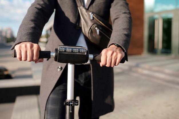 Handen van jonge elegante zakenman in vacht houden door zwarte handvatten van elektrische scooter tijdens het verplaatsen langs de weg in stedelijke omgeving