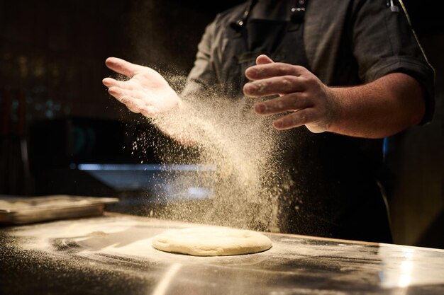 Handen van jonge mannelijke chef-kok die meel op deeg flatbread voor pizza strooit