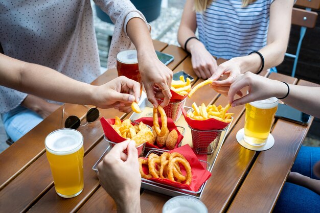 Handen van jonge mensen die wat gefrituurd voedsel nemen om te eten mix junkfood en bier op tafel snel en ongezond voedsel uienringen rustieke chips en frietjes