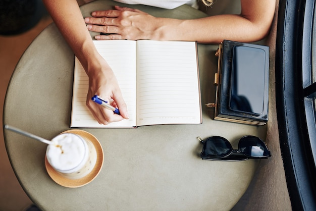 Foto handen van jonge vrouw cappuccino drinken en plannen schrijven voor de dag in planner, van bovenaf bekijken