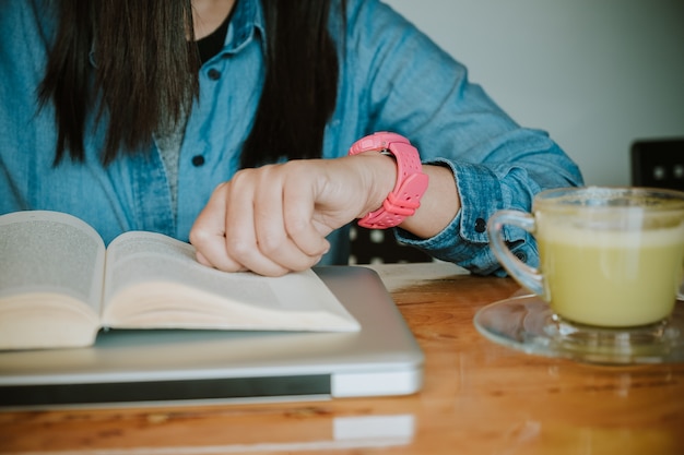 Handen van jonge vrouw die horloge bekijken terwijl het lezen van een boek op houten lijst.